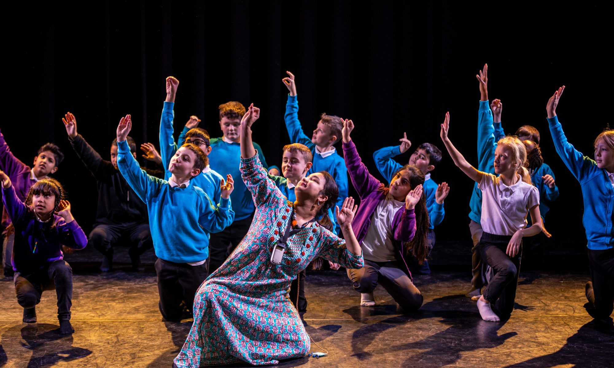 A woman leading a group of children in school uniforms in a dance, with her hand raised to the sky.