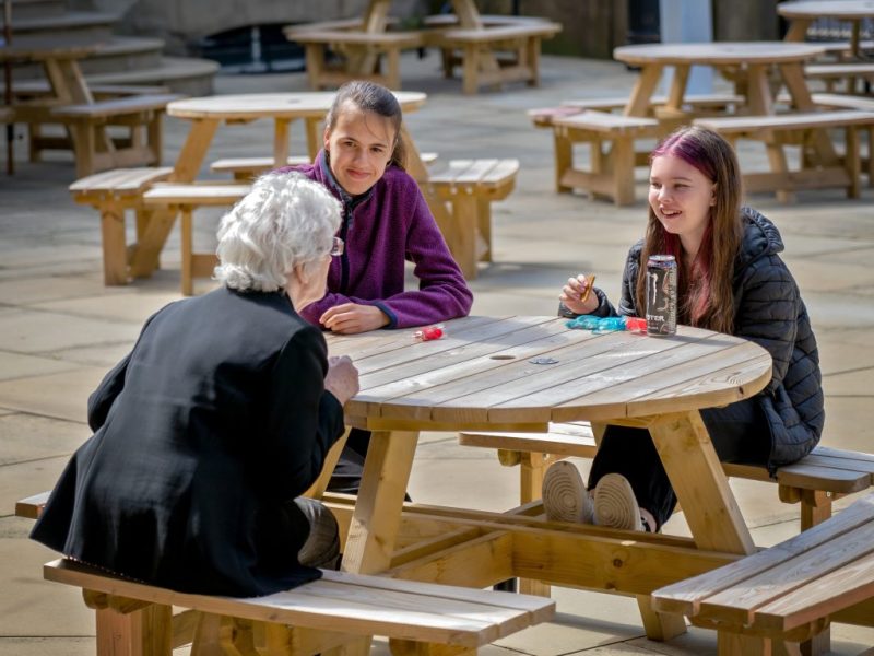 Older woman leaning across a bench to talk to two young people who are looking and smiling at her