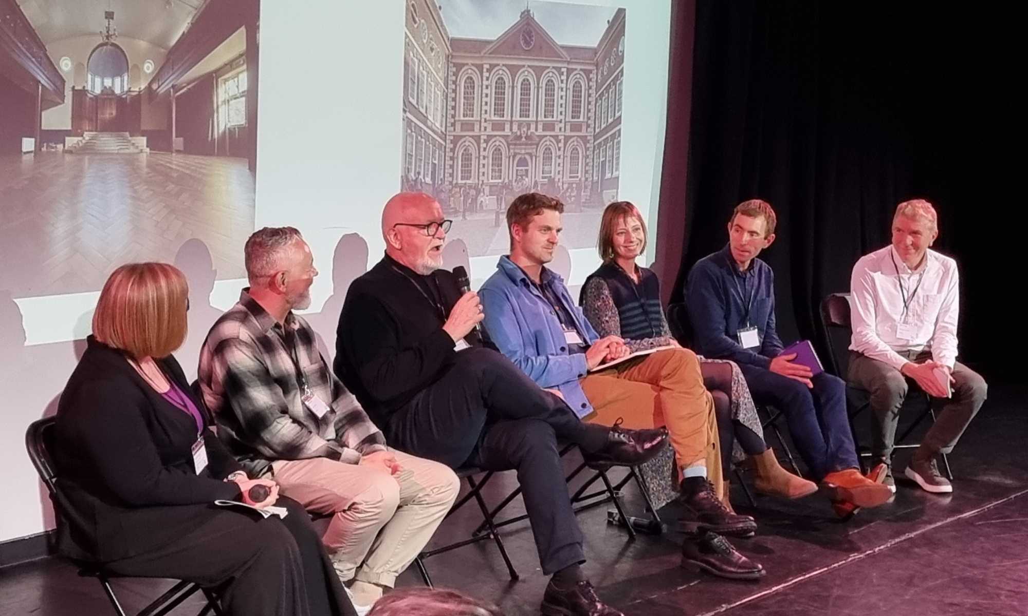 A panel of seven people sitting in front of a projection screen showing images of several Future Arts Centres venues