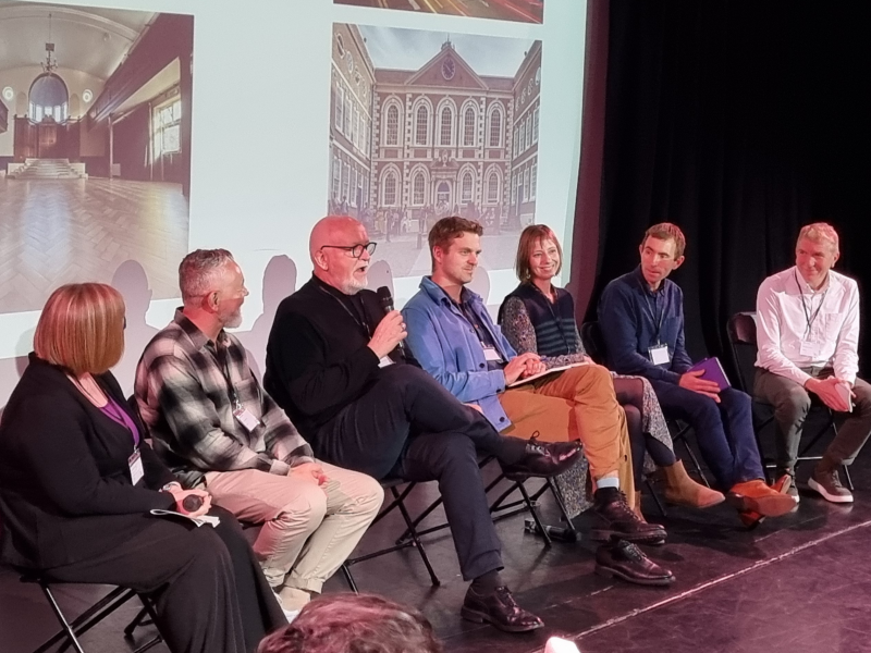 A panel of seven people sitting in front of a projection screen showing images of several Future Arts Centres venues