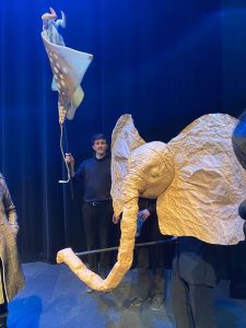 A performer with a puppet of a large, white elephant head