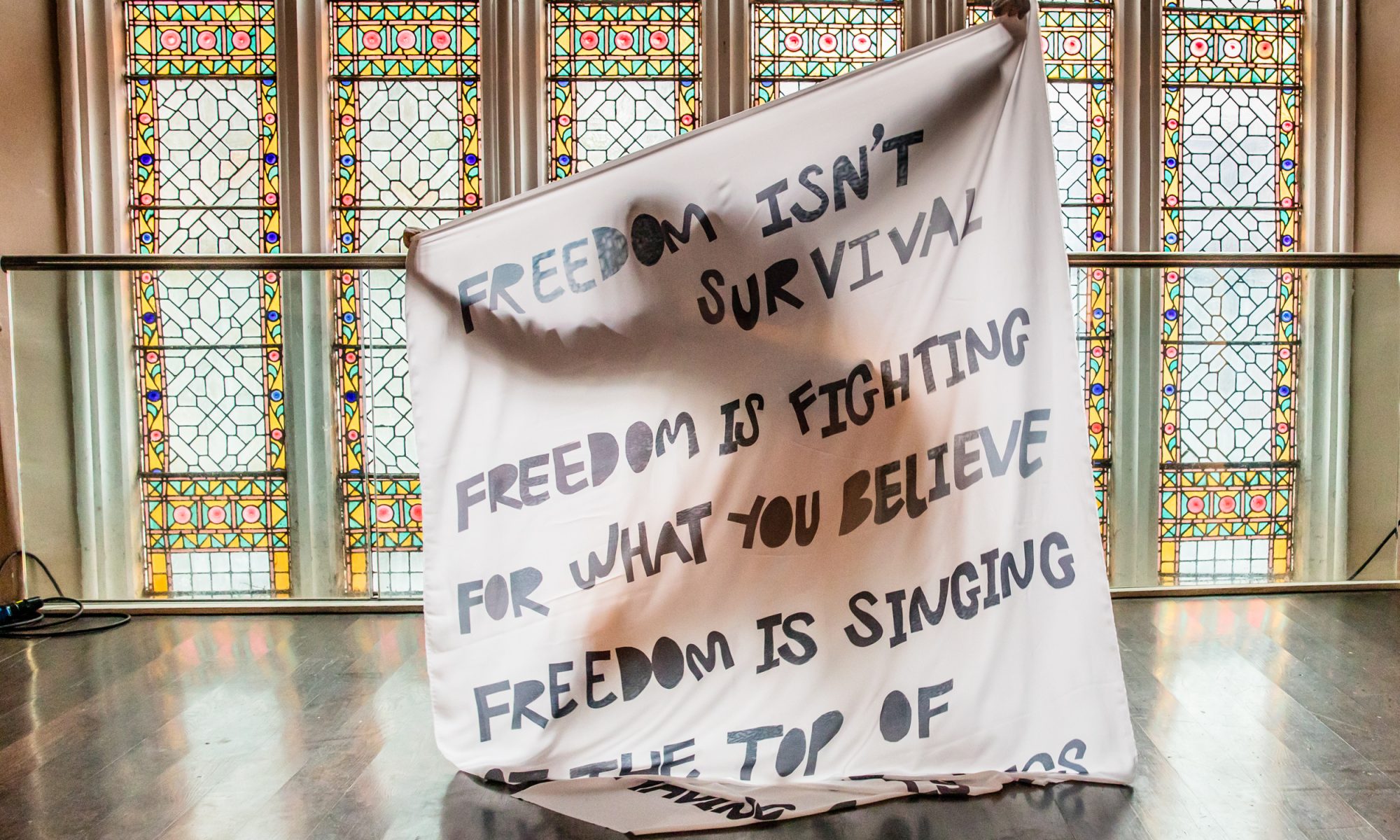 A person (hidden) holding up a large piece of material with text reading 'Freedom isn't survival, freedom is fighting for what you believe. Freedom is singing...' (rest of text not visible)