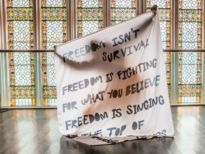 A person (hidden) holding up a large piece of material with text reading 'Freedom isn't survival, freedom is fighting for what you believe. Freedom is singing...' (rest of text not visible)