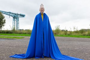 An older woman wearing a floor-length bright blue cape, and a mock crown