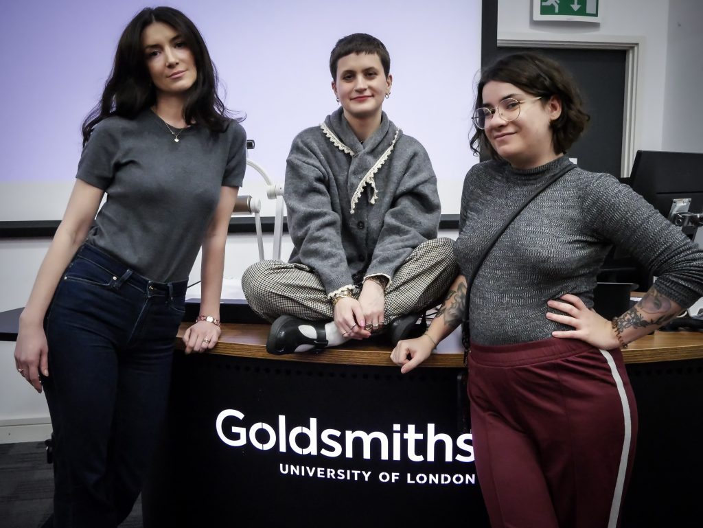 Three students face the camera. One is sitting cross-legged on a table that has the words 'Goldsmiths, University of London'