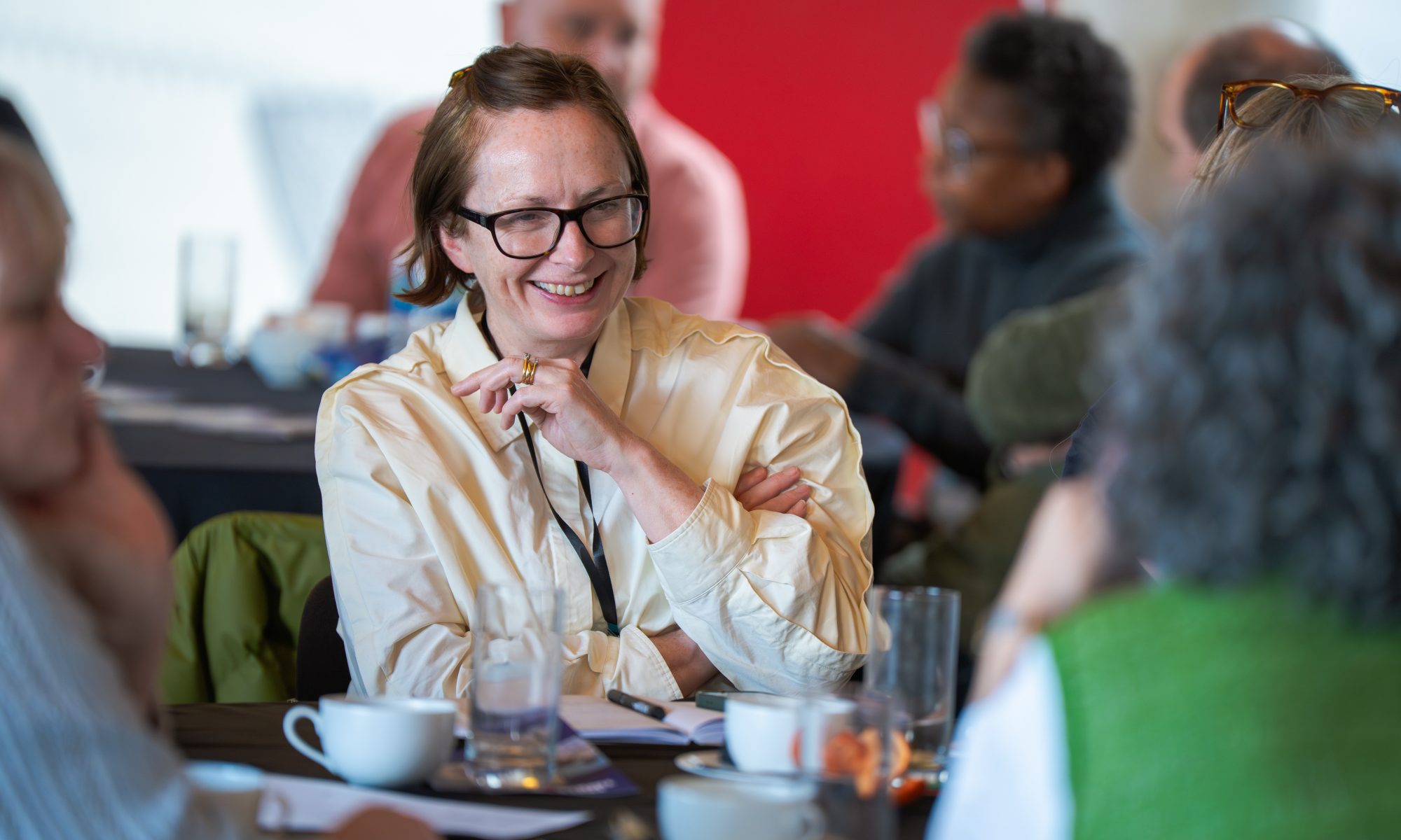 Members seated around tables and chatting at the Future Arts Centres Members' Meeting. The image focuses in one person with others blurred to either side and in the background.
