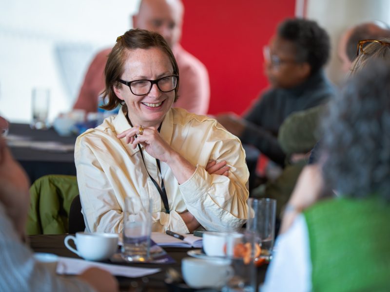 Members seated around tables and chatting at the Future Arts Centres Members' Meeting. The image focuses in one person with others blurred to either side and in the background.