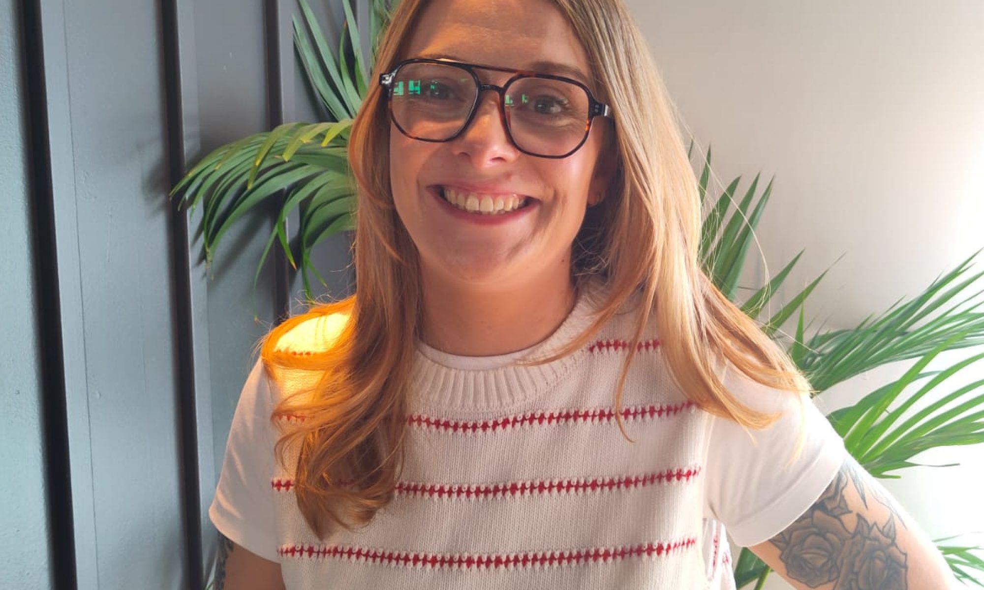 Elena Rowe smiles for the camera. She is wearing a white tank top with horizontal red stripes over a white t-shirt. She has long, fair hair and is wearing glasses.