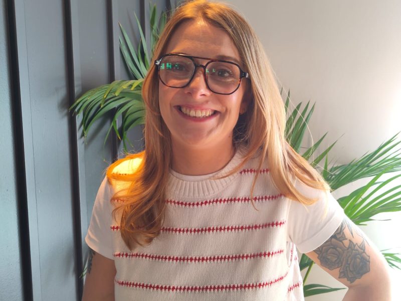 Elena Rowe smiles for the camera. She is wearing a white tank top with horizontal red stripes over a white t-shirt. She has long, fair hair and is wearing glasses.